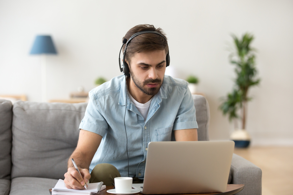 A man working from home on his laptop while wearing a headset with a mic and taking notes